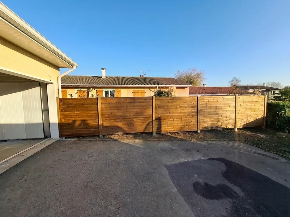 Clôture en bois horizontale récemment installée, séparant une allée goudronnée d'une propriété voisine